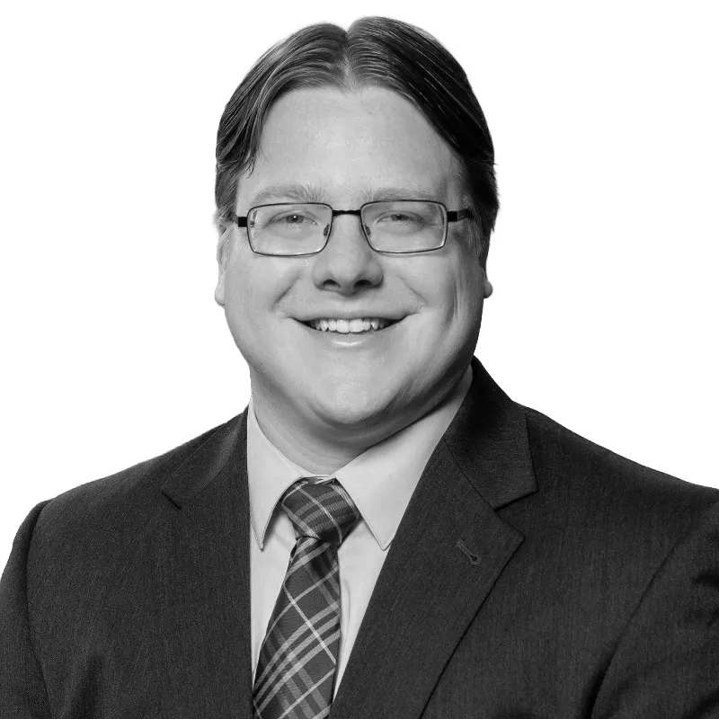 LUMITURE | Man with glasses wearing a suit, dress shirt, and plaid tie, smiling at the camera against a white background.